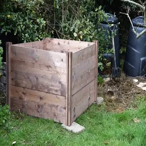 wooden garden compost bin in a grassy backyard beside shrubs and tools